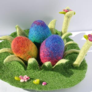 Könnte beinhalten: Ein Filznest mit drei bunten Ostereiern. Das Nest ist grün und hat zwei Filzblumen mit rosa Zentren. Die Eier sind blau, rot und lila.