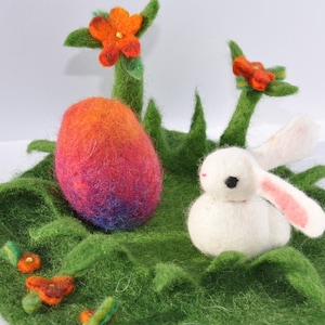 Puede incluir: Una escena de Pascua de fieltro con un huevo colorido, un conejito blanco con orejas rosas y flores naranjas. El huevo pasa de naranja a morado. El conejito está sobre una base de fieltro verde con hierba.