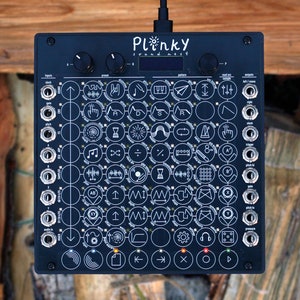 Puede incluir: Un sintetizador negro Plinky Sound-Kit con una cuadrícula de 48 botones y una pequeña pantalla LCD. Los botones tienen varios símbolos e iconos impresos en ellos.