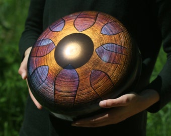 May include: A person holding a round, metallic handpan with a sun-like design. The handpan is decorated with intricate patterns in shades of purple, orange, and gold.