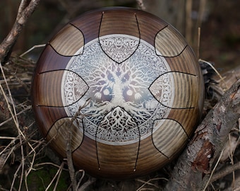 Può includere: Un handpan in legno marrone con un design ad albero della vita argentato. L'handpan è appoggiato su un letto di ramoscelli e rami.