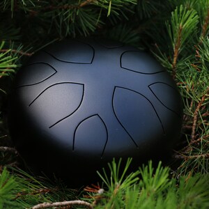 May include: A black steel handpan with a unique design of leaf-shaped indentations. The handpan is resting on a bed of green pine needles.