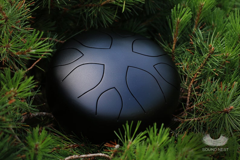 May include: A black steel handpan with a unique design of leaf-shaped indentations. The handpan is resting on a bed of green pine needles.