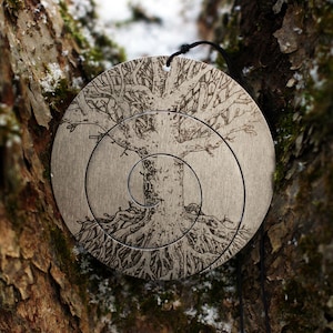 May include: A silver metal ornament with a tree design etched into the surface. The tree has a spiral pattern and is surrounded by concentric circles.