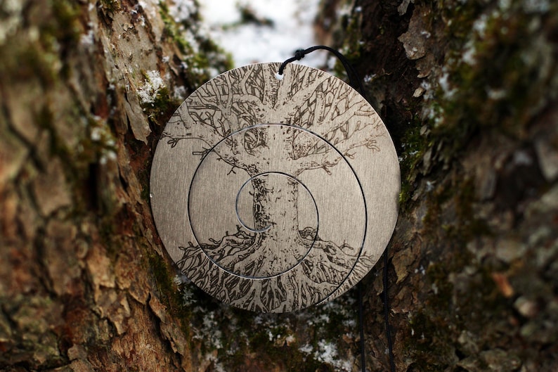 May include: A silver metal ornament with a tree design etched into the surface. The tree has a spiral pattern and is surrounded by concentric circles.
