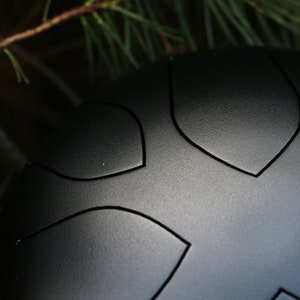 May include: A black steel tongue drum with a unique design featuring multiple indentations. The drum is made of steel and has a smooth, matte finish.