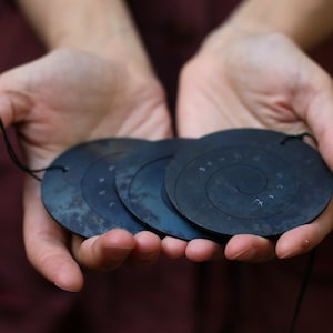 May include: Four black metal discs with a spiral design and numbers inscribed on them. The discs are strung together with a black cord.