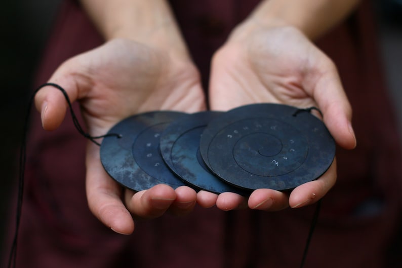 May include: Four black metal discs with a spiral design and numbers inscribed on them. The discs are strung together with a black cord.