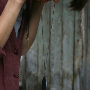 May include: A person wearing a maroon shirt with a black cord necklace and a black oval pendant.