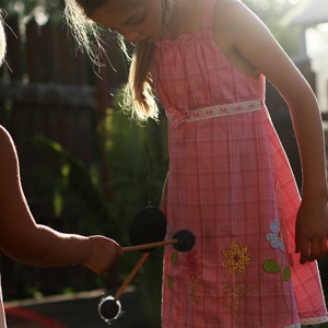 May include: Two young girls playing with a wooden toy with black discs. The girl in the pink dress is holding the toy and the girl in the white dress is holding the other end. The girls are outdoors in a backyard setting.