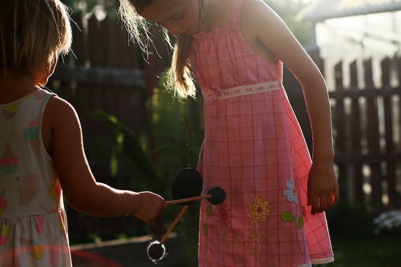 May include: Two young girls playing with a wooden toy with black discs. The girl in the pink dress is holding the toy and the girl in the white dress is holding the other end. The girls are outdoors in a backyard setting.