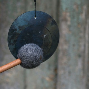May include: A black metal gong with a felt mallet. The gong has a circular shape and is hanging from a string. The mallet is attached to a wooden handle.
