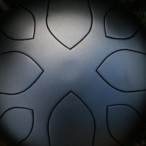 May include: A black steel handpan with a circular shape and a design of eight leaf-shaped indentations. The handpan is resting on a bed of green pine needles.