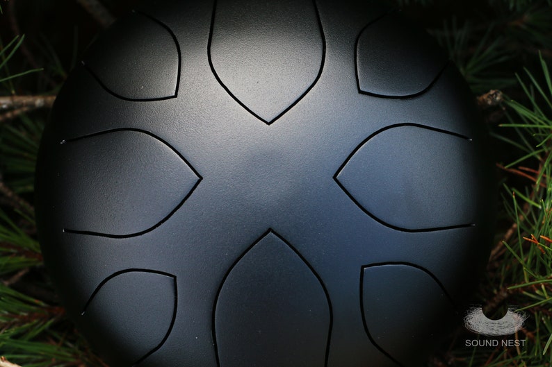 May include: A black steel handpan with a circular shape and a design of eight leaf-shaped indentations. The handpan is resting on a bed of green pine needles.