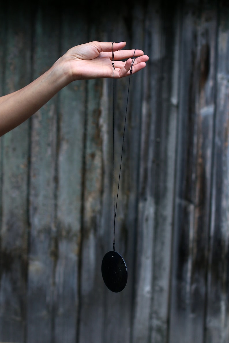 May include: A black disc pendant hanging from a black string held by a hand.
