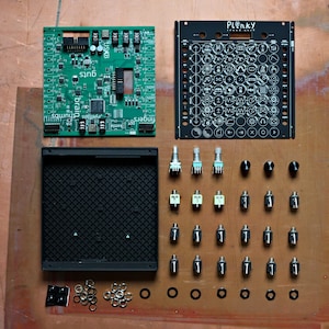 Puede incluir: Una vista cenital muestra componentes electrónicos para un proyecto de bricolaje. Se ve una placa de circuito verde con texto y un panel negro con símbolos. Varios mandos, conectores y piezas pequeñas están dispuestos sobre una superficie marrón.