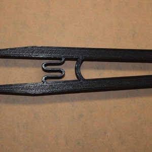 May include: A black 3D printed tool with a curved, spring-like mechanism in the center. Two white round objects are held in place by the tool.