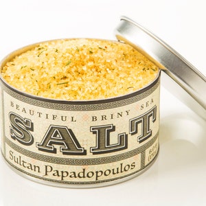 May include: A round metal tin filled with yellow sea salt. The label reads "Beautiful Briny Sea Salt" and "Sultan Papadopoulos".