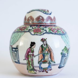 Può includere: Un vaso di porcellana bianca con un coperchio, decorato con un motivo floreale colorato e una scena di due figure in abiti tradizionali cinesi. Il vaso ha un corpo rotondo e un collo stretto.