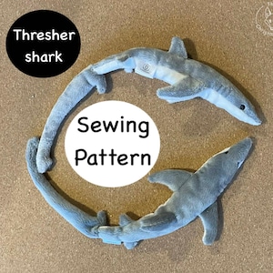 May include: Two plush thresher shark toys in a circular arrangement. The sharks are gray with white bellies and have long tails. The text "Thresher shark" is in a black circle, and "Sewing Pattern" is in a white circle.