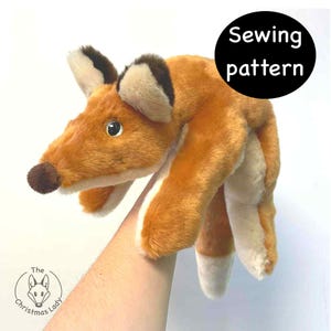 Könnte beinhalten: Eine Fuchs-Handpuppe aus Plüsch mit orangefarbenem und weißem Fell, braunen Akzenten und einer dunkelbraunen Nase. Die Puppe wird von einem Arm gehalten. Ein schwarzer Kreis mit den Worten "Sewing pattern" befindet sich im Hintergrund.