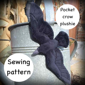 Op de afbeelding: Een donkergrijze pluche kraai met uitgespreide vleugels, rustend op een metalen gieter. De kraai heeft een zacht, getextureerd uiterlijk. Tekst op de afbeelding luidt "Pocket crow plushie" en "Sewing pattern".