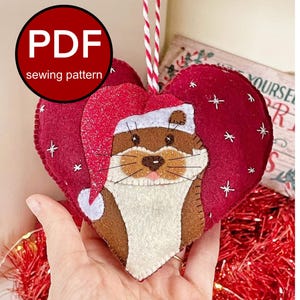 Puede incluir: Adorno navideño en forma de corazón con una nutria de fieltro con un gorro de Papá Noel. El adorno es rojo con detalles de copos de nieve blancos y una cuerda blanca y roja para colgar. El texto "PDF sewing pattern" está en un círculo rojo.