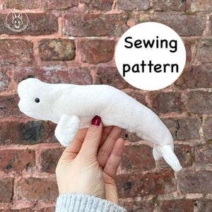 May include: A white plush beluga whale toy with small fins and a tail. The toy has a black eye and is held against a brick wall. A white circle with the words "Sewing pattern" is in the background.