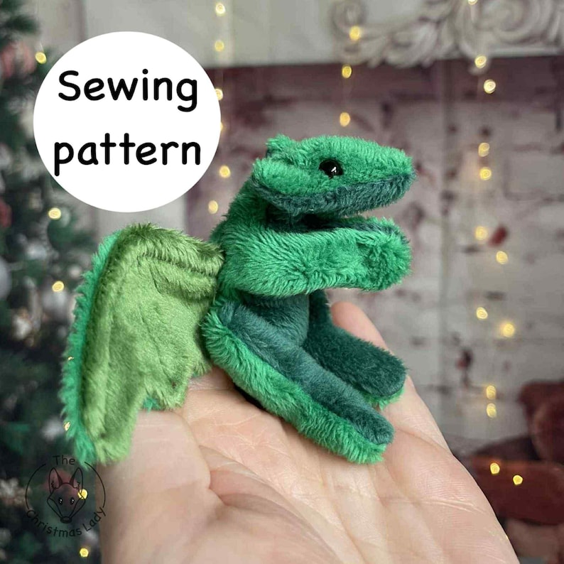 Puede incluir: Un juguete de drag&oacute;n verde con alas, sostenido en una mano. El drag&oacute;n est&aacute; hecho de tela suave y texturizada. El texto "Sewing pattern" est&aacute; en un c&iacute;rculo blanco en el fondo.