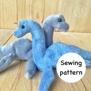 Può includere: Due dinosauri di peluche blu e grigi, con occhi neri e colli lunghi. I giocattoli sono su una superficie di legno. Un cerchio bianco con le parole "Sewing pattern" è nell'immagine.