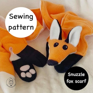 Può includere: Sciarpa a forma di volpe arancione e bianca con dettagli neri. La sciarpa presenta una testa di volpe con orecchie e occhi neri. Le zampe sono nere con cuscinetti bianchi. I testi "Sewing pattern" e "Snuzzle fox scarf" sono visibili.