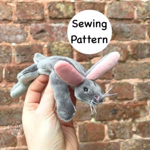 May include: A gray plush bunny with pink inner ears and a pink nose is held in a hand. The bunny has white whiskers and black eyes. The words "Sewing Pattern" are in a white circle above the bunny.