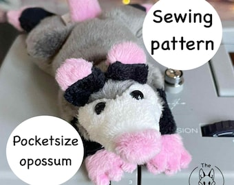 Könnte beinhalten: Ein kleines, plüschiges Opossum-Spielzeug mit grauem Fell, rosa Pfoten und schwarzen Akzenten. Der Text "Pocketsize opossum" und "Sewing pattern" sind im Bild zu sehen. Das Logo The Christmas Lady ist ebenfalls sichtbar.