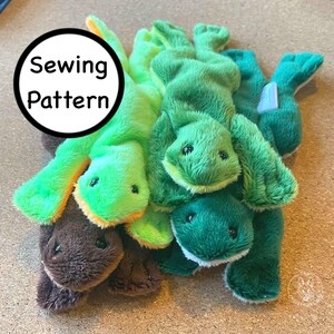 May include: A pile of plush toy lizards in shades of green and brown. The toys have small black eyes and are made of soft, fuzzy material. A white circle with the words "Sewing Pattern" is in the upper left corner.