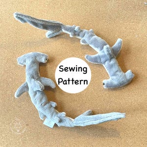 May include: Two gray plush hammerhead sharks are displayed on a light brown surface. The sharks have a soft, textured appearance with detailed fins and tails. A white circle with the words "Sewing Pattern" is in the center.