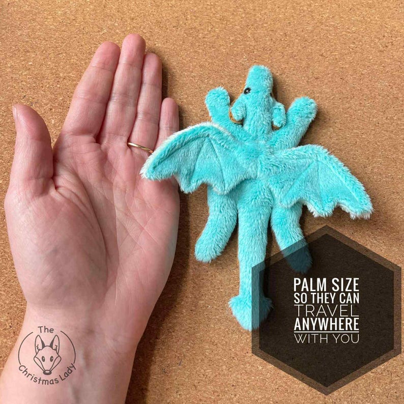 Puede incluir: Un juguete de peluche de drag&oacute;n turquesa con alas extendidas y una larga cola. El drag&oacute;n se muestra junto a una mano, con el texto "PALM SIZE SO THEY CAN TRAVEL ANYWHERE WITH YOU". El drag&oacute;n tiene peque&ntilde;os ojos negros y una nariz marr&oacute;n.