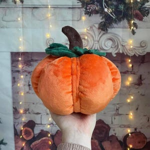May include: A plush orange pumpkin toy with a brown stalk and green leaves. The pumpkin is round and has vertical ridges. A hand is holding the pumpkin.