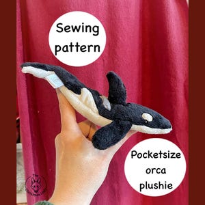 May include: A black and white orca plushie with a cream-colored belly is held up. The plushie has a small fin and a white spot near its eye. Text on the image reads "Sewing pattern" and "Pocketsize orca plushie."