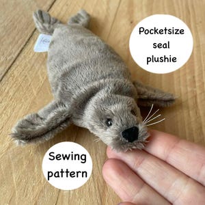Può includere: Un giocattolo di peluche grigio a forma di foca con naso nero e baffi bianchi. I testi "Pocketsize seal plushie" e "Sewing pattern" sono in cerchi bianchi. La foca è su una superficie di legno.