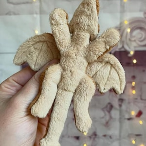 Puede incluir: Un juguete de drag&oacute;n de peluche beige con detalles marrones en las alas, la cabeza y los pies. El drag&oacute;n tiene cuatro patas y dos alas, y est&aacute; hecho de un material suave y difuso. El juguete est&aacute; sostenido en una mano.