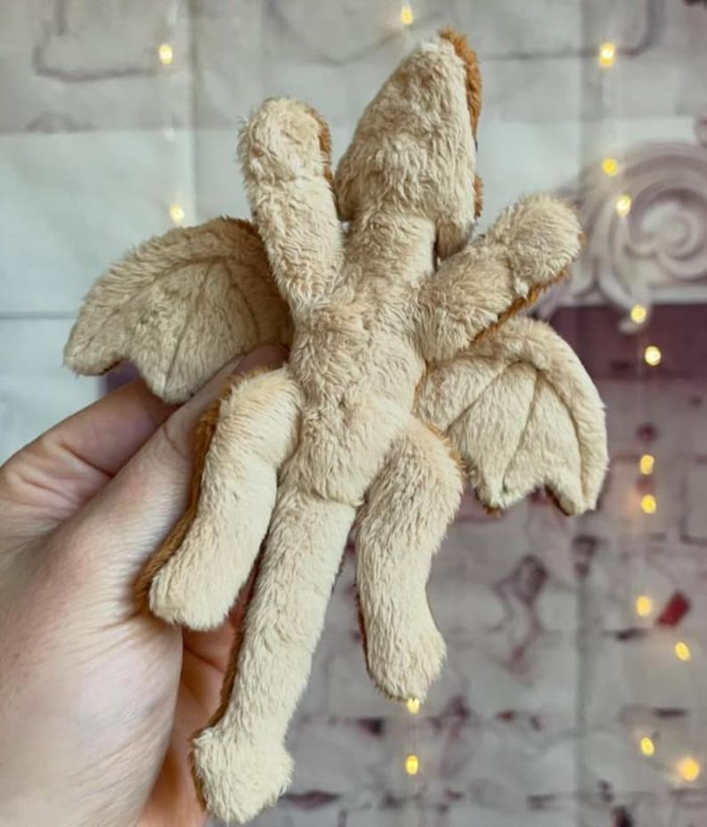 Puede incluir: Un juguete de drag&oacute;n de peluche beige con detalles marrones en las alas, la cabeza y los pies. El drag&oacute;n tiene cuatro patas y dos alas, y est&aacute; hecho de un material suave y difuso. El juguete est&aacute; sostenido en una mano.