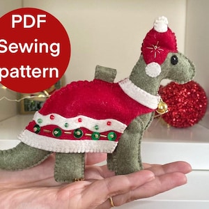 May include: A felt dinosaur ornament wearing a red Santa suit and hat, with a gold bell. The ornament has the text "PDF Sewing pattern" on a red circle. A festive holiday decoration.