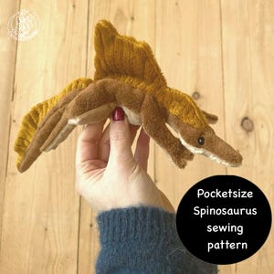 Op de afbeelding: Een bruin en bruin Spinosaurus dinosaurus knuffel met een opvallende zeil op zijn rug. Het speelgoed wordt in een hand gehouden, en een zwarte cirkel met witte tekst die luidt "Pocketsize Spinosaurus sewing pattern" staat in de afbeelding.
