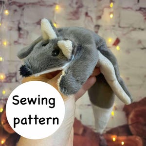 May include: A plush gray and white toy animal with a black nose and ears. The text "Sewing pattern" is in a white circle below the toy. The toy is held in a hand.