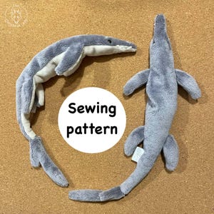 May include: Two plush, gray mosasaur toys with white bellies are displayed. The toys have flipper-like appendages and long tails. A white circle with the words "Sewing pattern" is in the center of the image. The toys are on a brown surface.