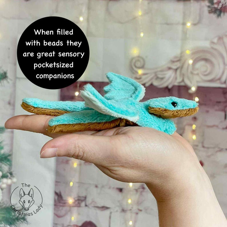 Puede incluir: Un drag&oacute;n de peluche turquesa con detalles marrones y alas blancas. El drag&oacute;n se sostiene en una mano. El texto en un c&iacute;rculo negro dice: "Cuando se llenan de cuentas, son grandes compa&ntilde;eros sensoriales de bolsillo".