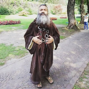 May include: A man with a long gray beard is wearing a brown robe with a chain necklace. He is standing on a brick pathway in a park.