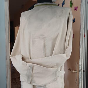 May include: A beige canvas straightjacket featuring long sleeves that wrap around the front and secure with straps. The garment has a high collar with a subtle olive green trim and a reinforced rectangular panel on the chest area, displayed on a mannequin.