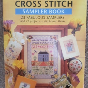 May include: A book titled "The New Cross Stitch Sampler Book" with a yellow and blue cover. The cover features a cross-stitch sampler with a house, flowers, and text. Additional cross-stitch projects and accessories are displayed around the book.