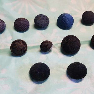 May include: A collection of 12 round buttons in various shades of blue and black. The buttons are made of fabric and have a textured surface.